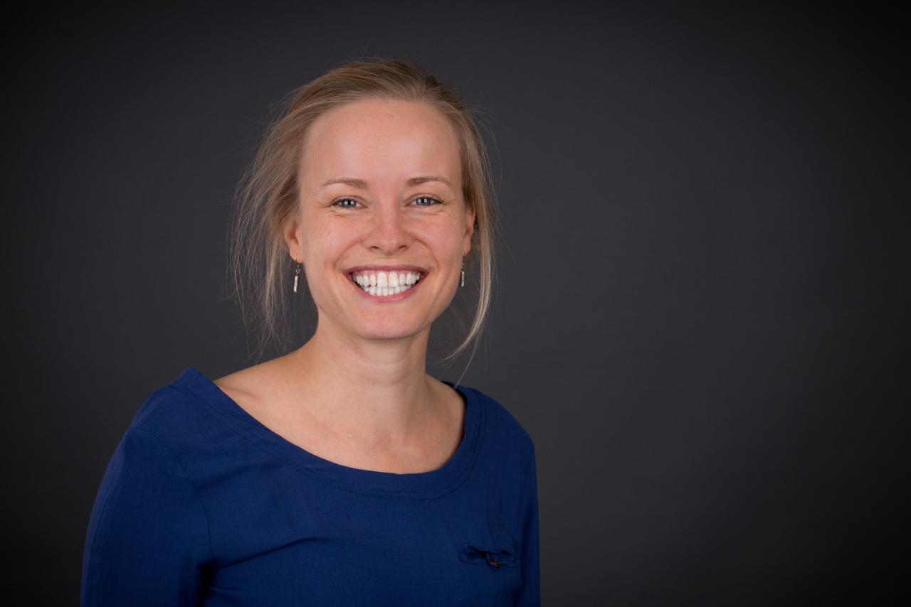 Maria Müller lächelt in die Kamera. Sie ist vor braunem Hintergrund halblinks im Bild zu sehen mit blauem Oberteil und mittellangen, zurückgebundenen blonden Haaren.